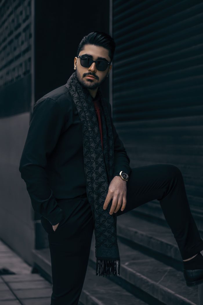 Man in stylish attire posing confidently outdoors with a dark scarf and sunglasses.