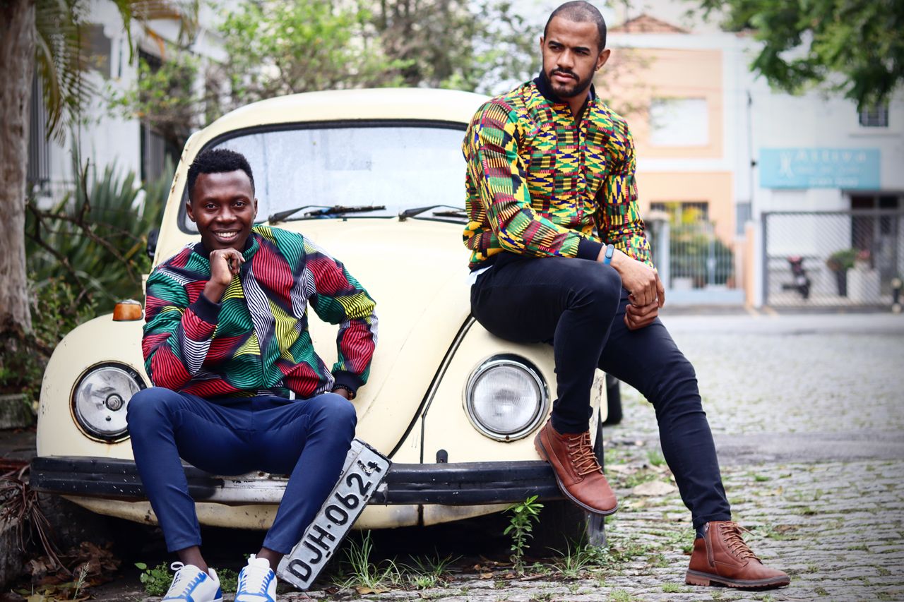 Crafting Captivating Headlines: Your awesome post title goes here Two men wearing vibrant African print shirts posed with a classic yellow Volkswagen Beetle outdoors.